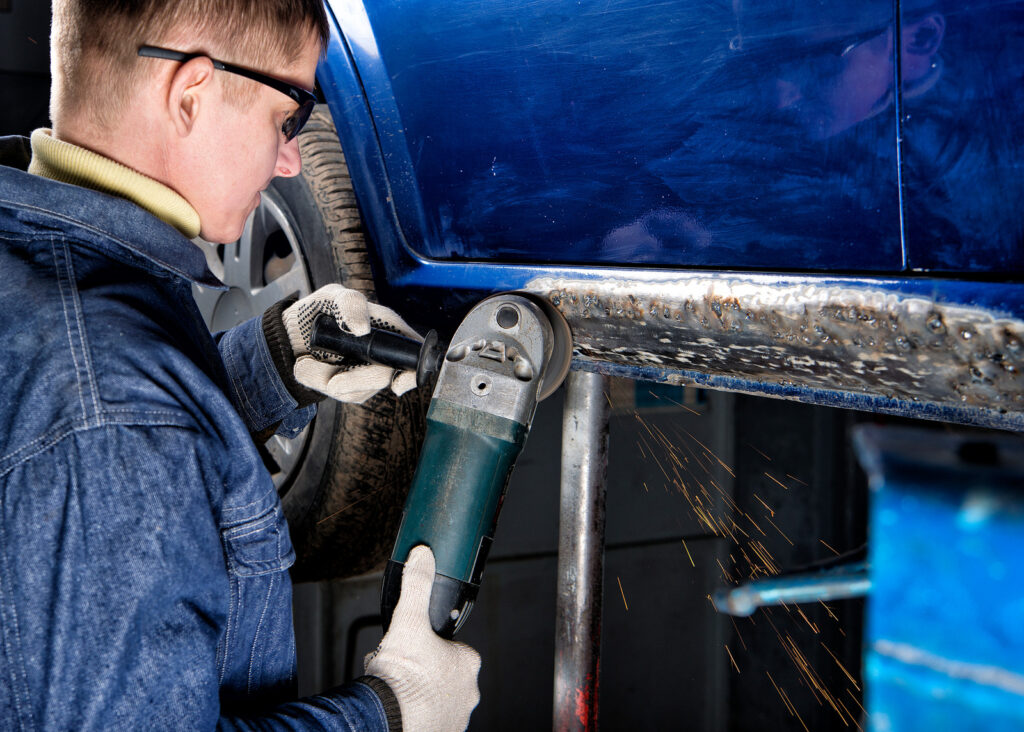 Un trabajador utilizando una herramienta eléctrica para pulir la parte inferior de un automóvil azul elevado, con chispas volando mientras trabaja.