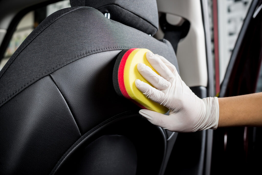 Una mano con guantes de látex limpiando el respaldo de un asiento de coche con una esponja, destacando la limpieza detallada del interior del vehículo.