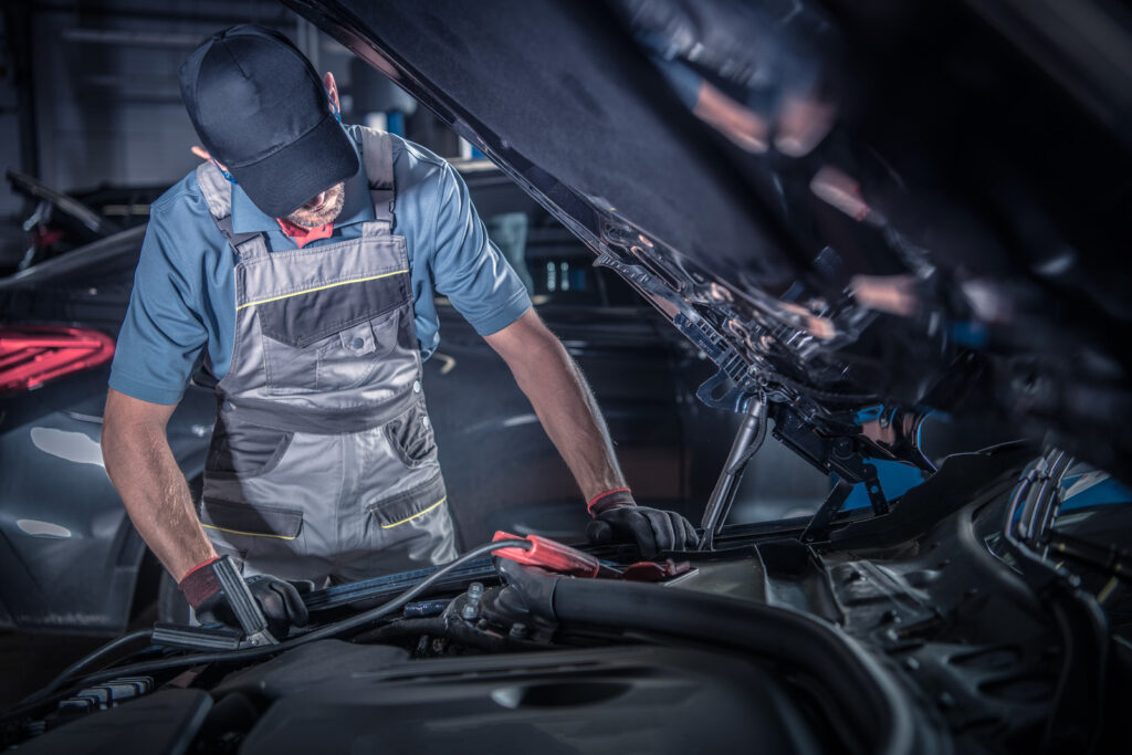 Un mecánico con gorra y overol trabajando bajo el capó levantado de un automóvil en un taller.