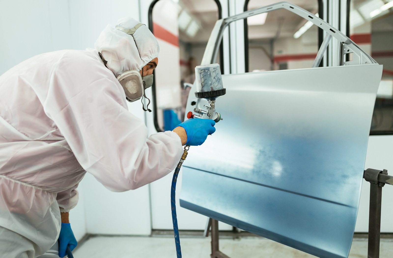 Trabajador en traje de protección y mascarilla, utilizando una pistola de pulverización para pintar una puerta de automóvil en un taller de pintura.