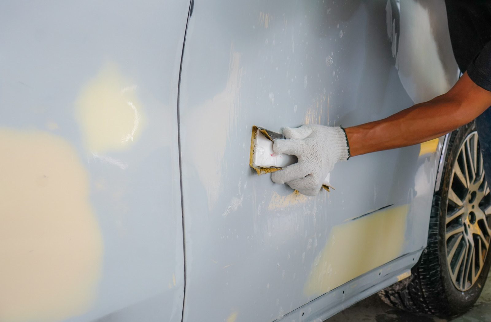 Primer plano de una mano con guante lijando la puerta de un automóvil con masilla aplicada, preparándola para pintar.